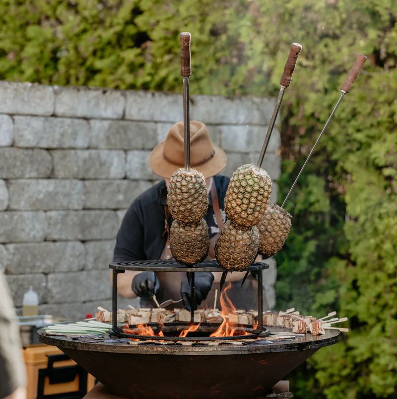 Ananas an Spießen über der offenen OFYR-Feuerplatte mit einem EMBER-Koch im Hintergrund